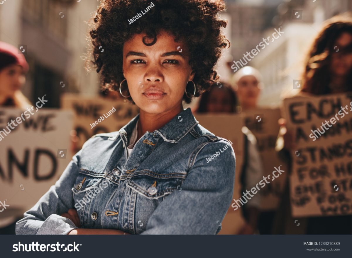 Young black woman with a group of demonstrators in the background outdoors. African woman protesting with a group of activists outdoors on the road.
